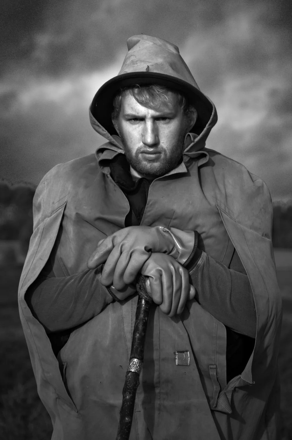 Black and white portrait of a shepherd in Northern Germany, with his hands folded on the top of a crook, set against a dark and stormy sky