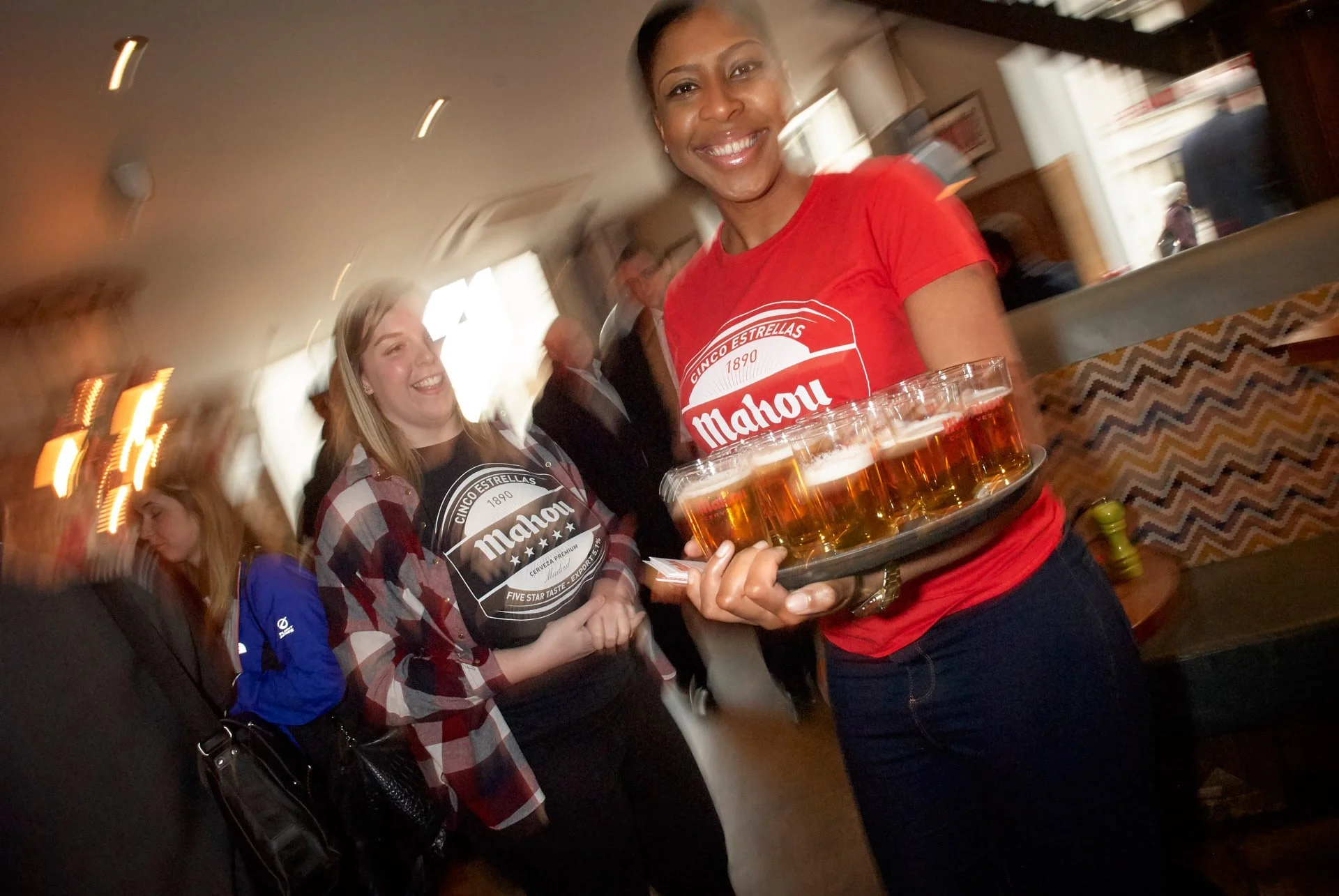 Smiling waitresses are carrying trays with beer in a busy and lively bar