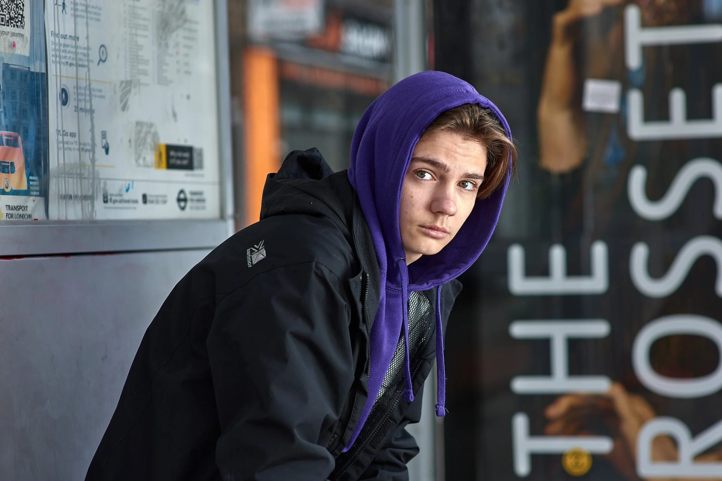A young man wearing a purple hooded top is sitting in a bus shelter, leaning forward and looking straight at the camera. Shot for the youth homelessness charity Centrepoint.