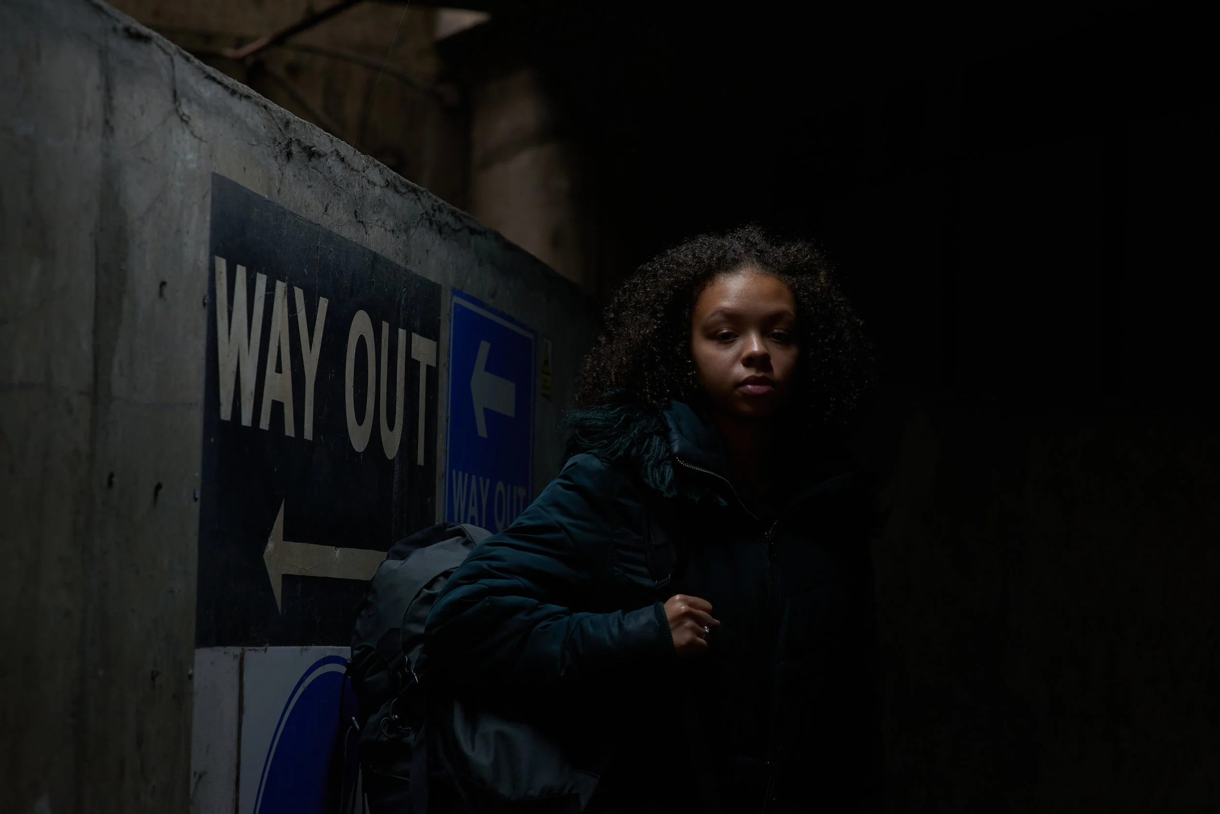 A vulnerable young woman is standing in the entrance to an underground car park in a dimly lit and threatening looking environment, shot as part of a fundraising campaign for the youth homelessness charity Centrepoint