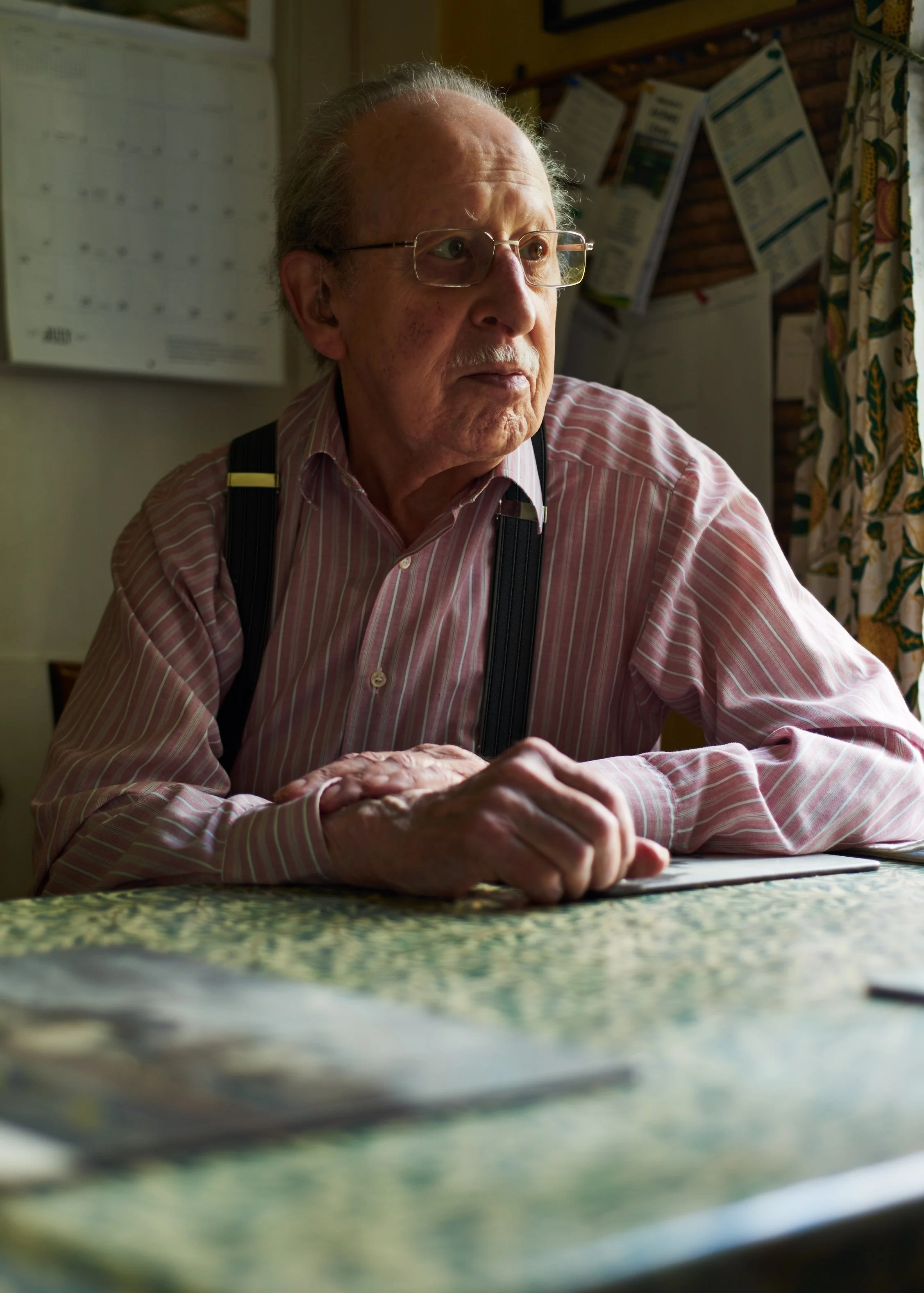 Portrait of an elderly man with glasses wearing a striped shirt and braces sitting at his kitchen table with light from a window coming in from one side