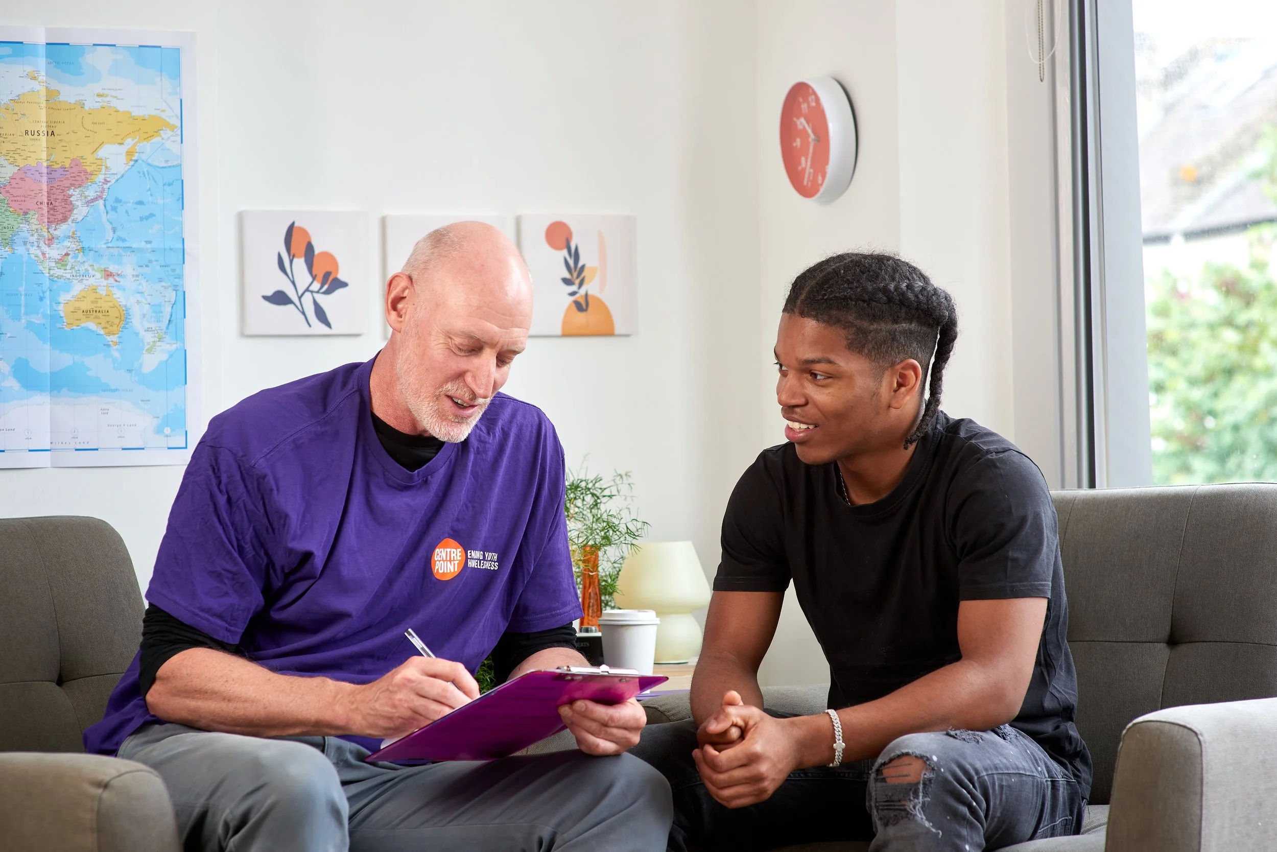 Portrait of a man wearing a purple T shirt with the Centrepoint logo smiling as he talks to a younger man sitting next to him