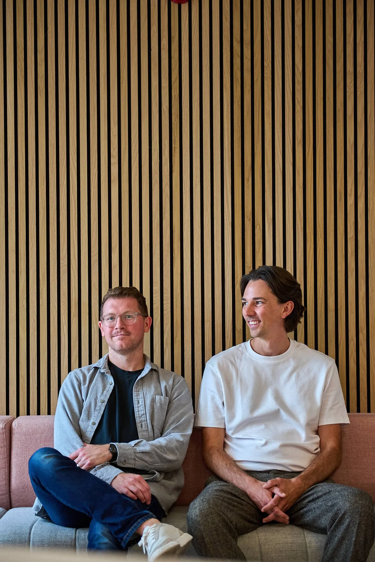 Portrait of two casually dressed men sitting on a sofa in front of a plain background of vertical wooden strips. One man is looking to the camera left and the other is facing the camera. Both are smiling