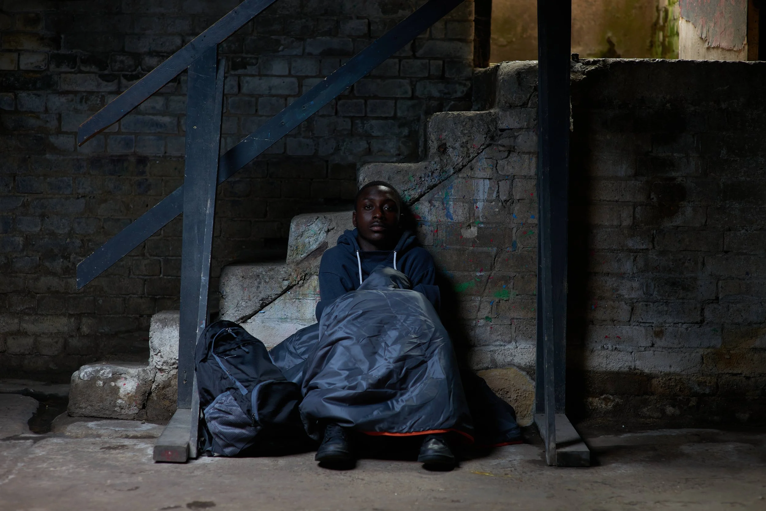 A young man is sitting at the bottom of a small set to steps in a dark and threatening environment. Shot as part of an awareness campaign for the youth homelessness charity Centrepoint
