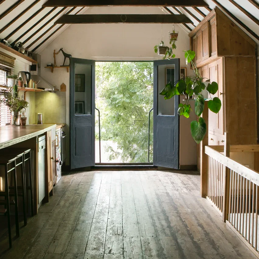 Bright kitchen with open doors leading to a tree-filled view outside, featuring wooden floors, shelves with kitchen items, and hanging plants.