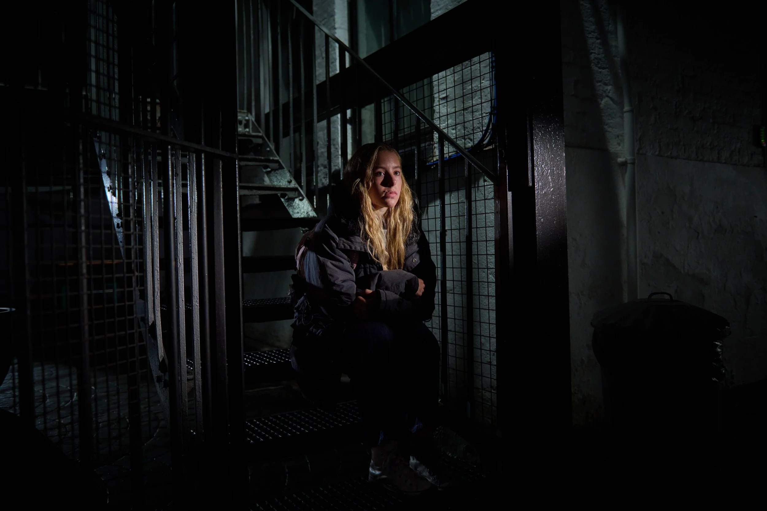 Portrait of a vulnerable looking young blonde haired woman sitting on an industrial looking staircase in the dark