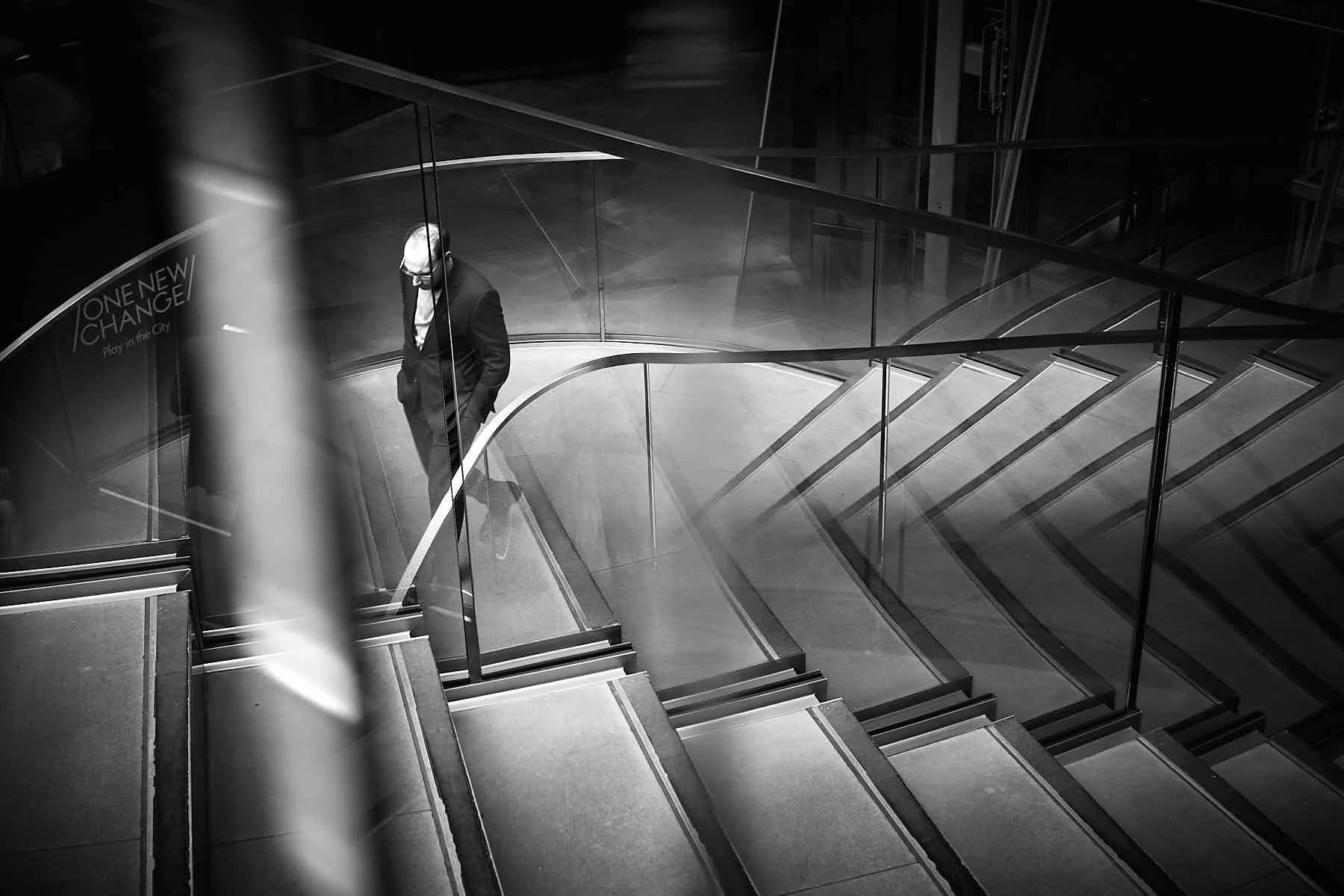 Black and white image of a man walking up a curved staircase with glass reflections all around