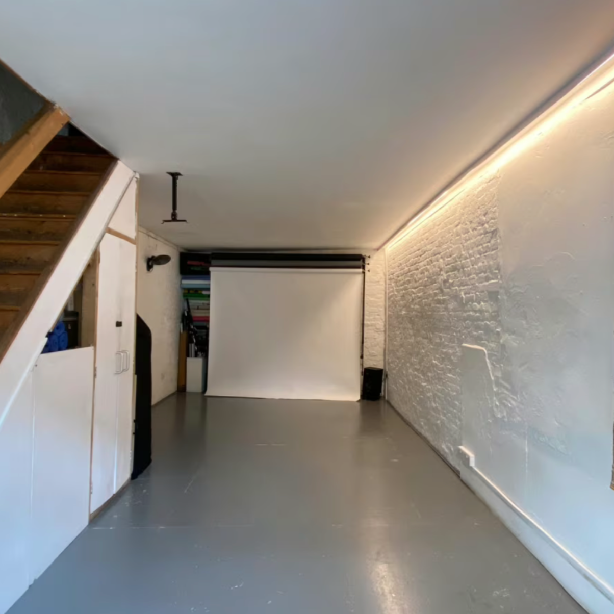 Empty room with white brick wall, gray floor, and a white projection screen. There is a wooden staircase on the left, a small cabinet with shelves, and a black speaker on the floor. Ambient lighting runs along the ceiling edge.