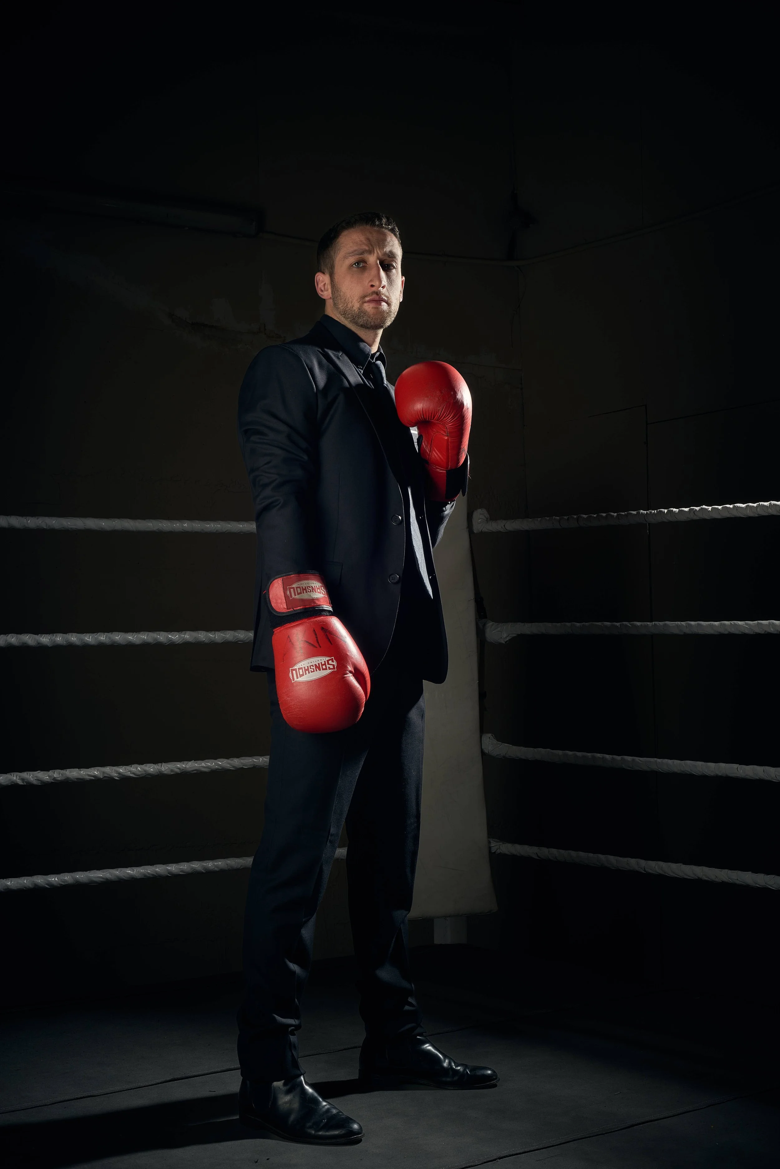 Full length portrait of a young man wearing a smart suit and boxing gloves, standing in a the corner of a boxing ring in a dark and moody atmosphere