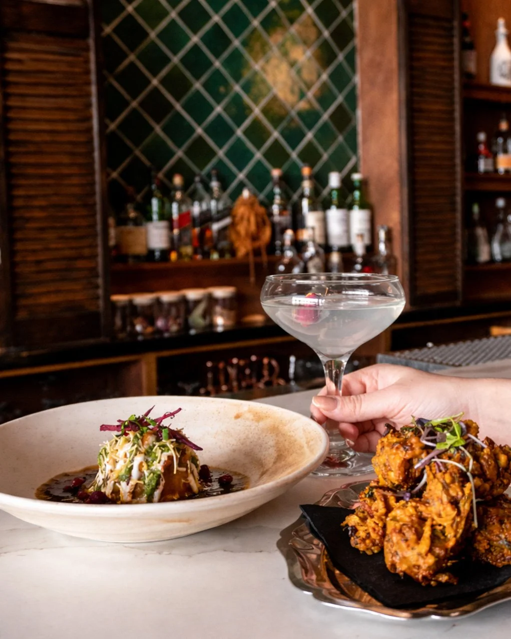A table with a bowl of salad topped with microgreens, a plate of fried chicken, and a cocktail in a coupe glass being held by a person. In the background, a bar with liquor bottles and a green decorative wall.