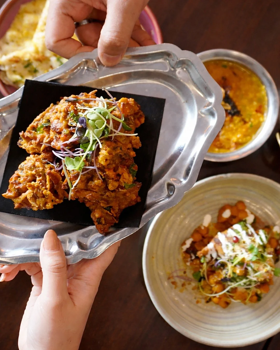 Hand holding a metal tray with a serving of Indian curry topped with microgreens, with bowls of curry and chickpea salad in the background.