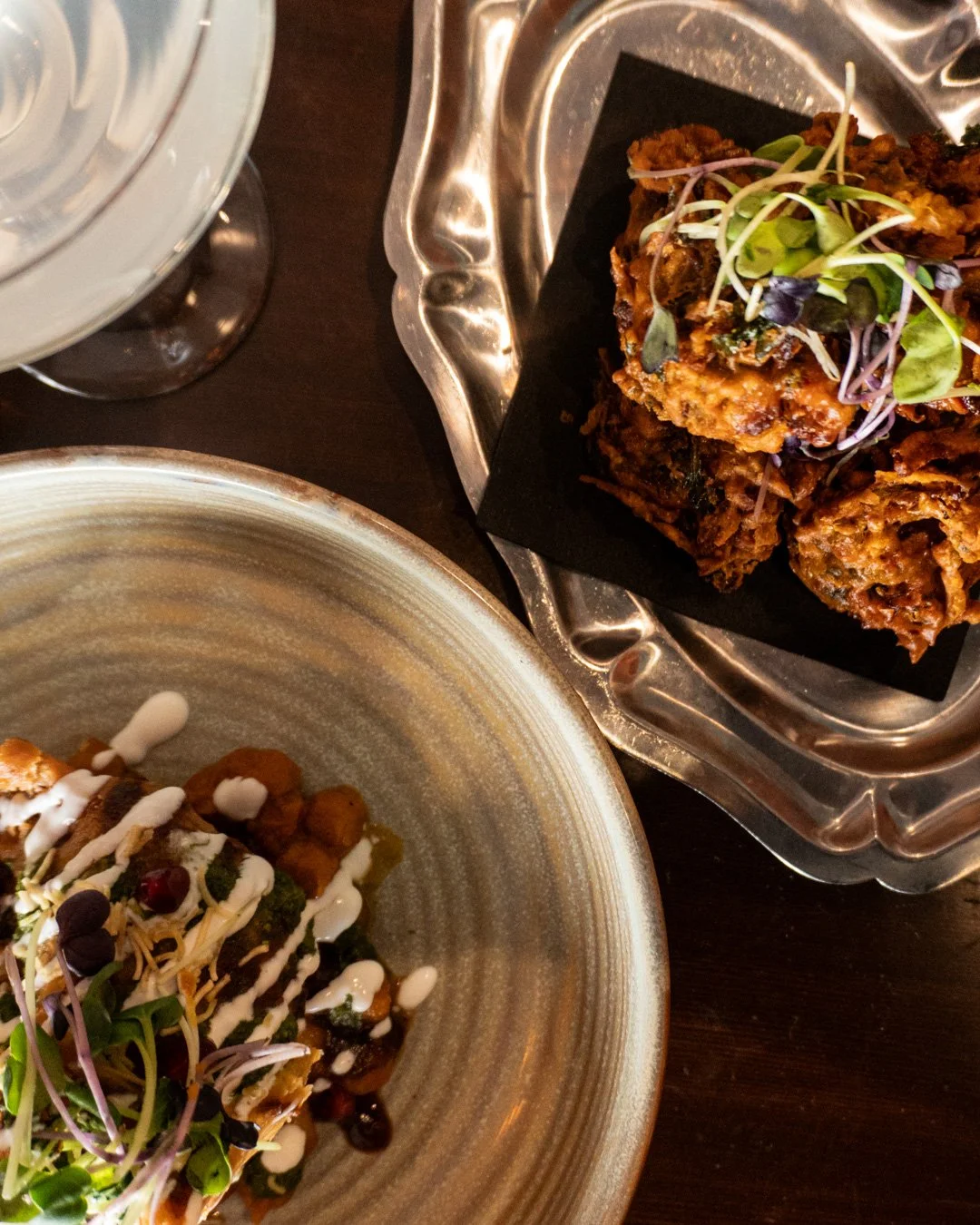 Close-up of two plates with baked dishes, garnished with microgreens, on a dark wooden table, with a glass of water nearby.
