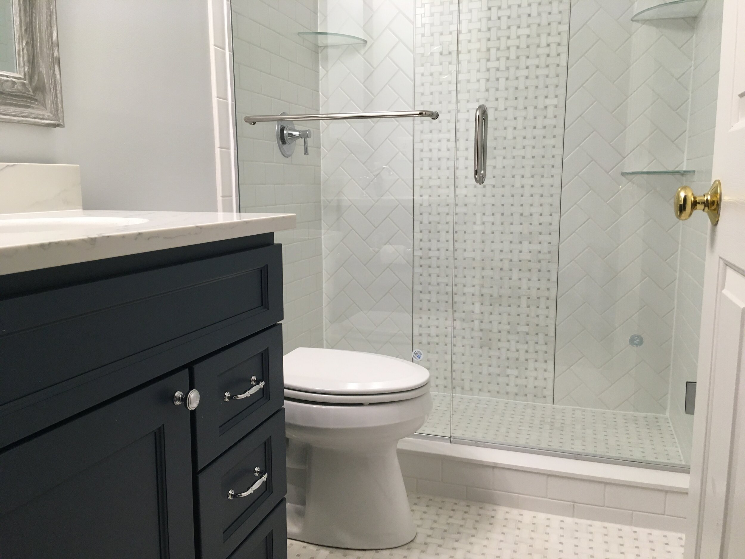  A finished bathroom with 3/8 thick glass door and fixed panel with the raincoat coating . The floor is Althea Glacier white basket weave with grey dots The same tile was used on the center of the back wall and white tile installed in herringbone des