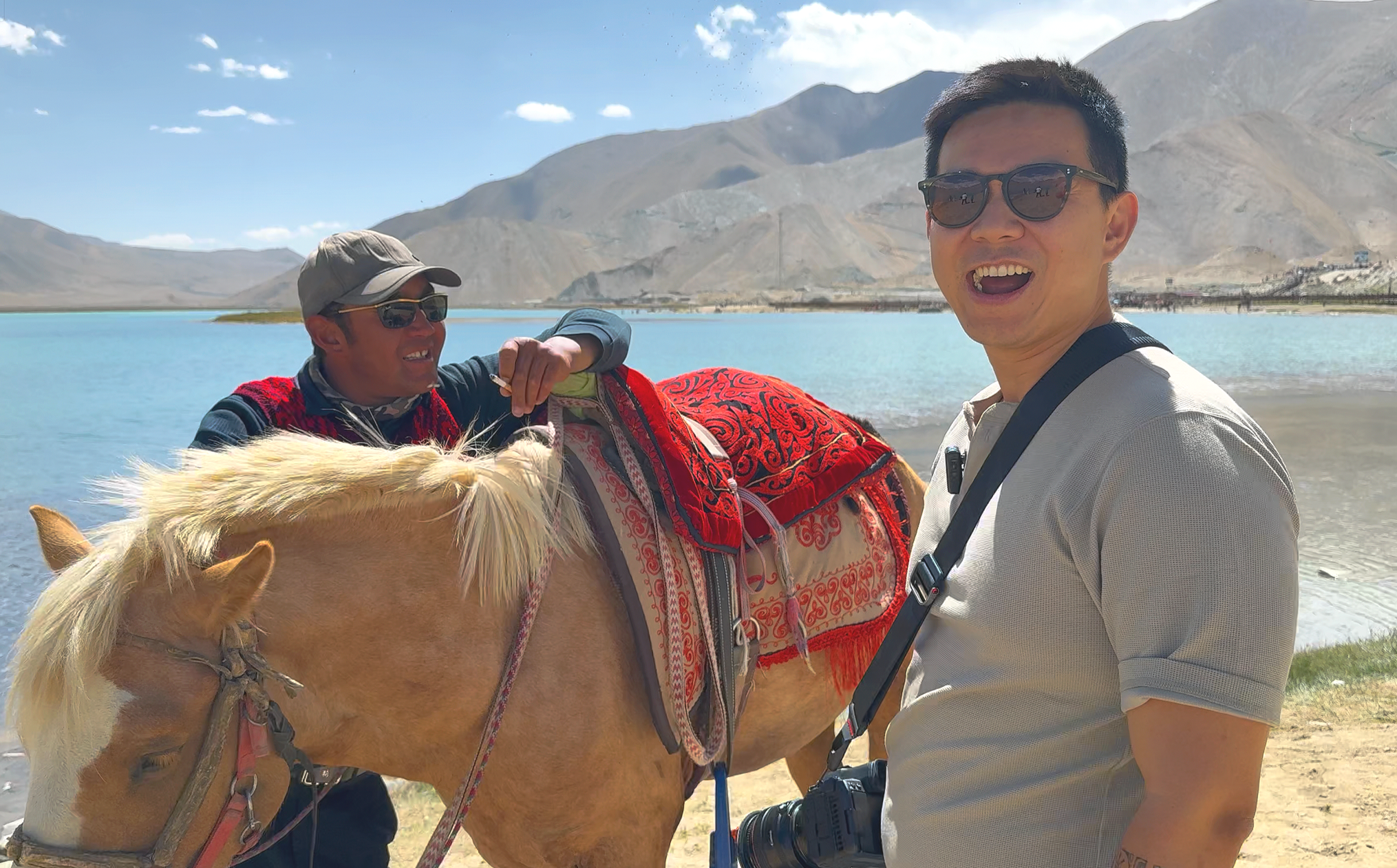 A man with sunglasses smiling at the camera stands next to a horse wearing decorative red saddle blankets. A person on the horse adjusts the saddle, with a lake and mountains in the background under a blue sky.