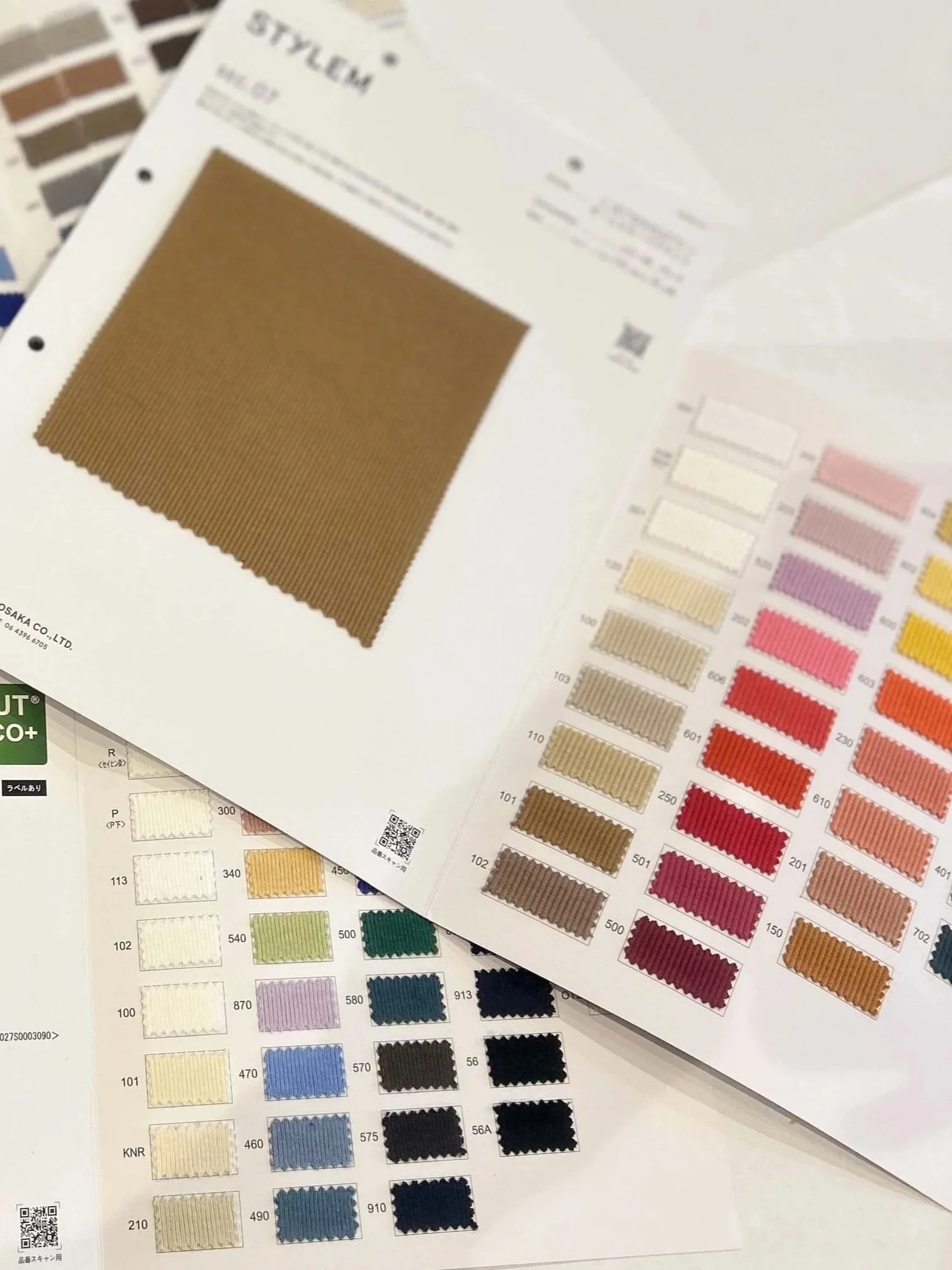 Close-up of fabric sample swatches arranged on printed color and fabric sample charts, with additional fabric swatches partially visible in the background.