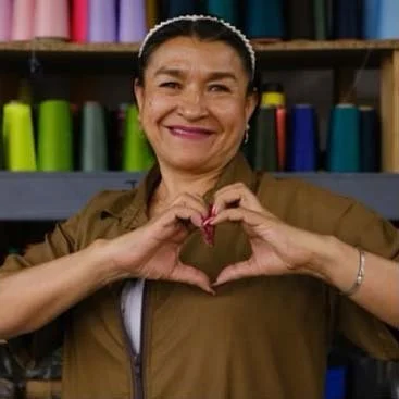 A woman smiling and making a heart shape with her hands in front of a colorful backdrop of fabric rolls.