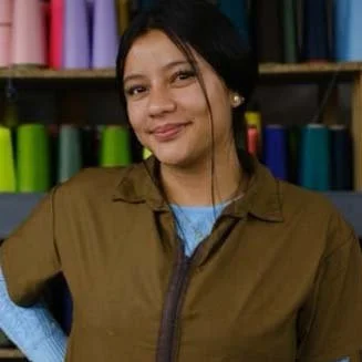 A woman smiling in front of a background of colorful fabric rolls in a textile shop.