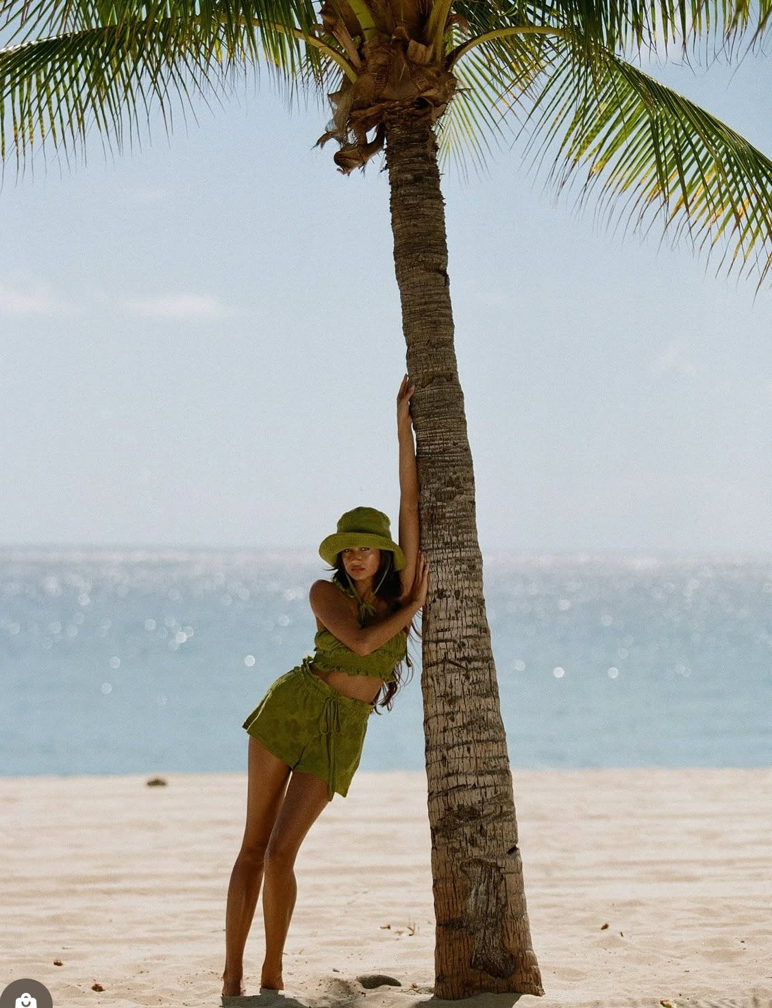 A woman in a green outfit and hat hugging a palm tree on a sandy beach with the ocean in the background.