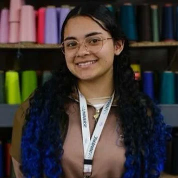 Young woman with glasses and long, curly hair smiling, standing in front of colorful yarn spools.