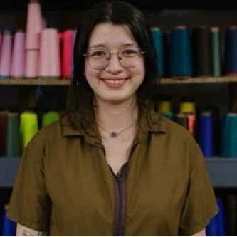 A young woman with glasses and shoulder-length dark hair, smiling, standing in front of shelves with colorful yarn spools.