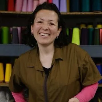 A smiling woman with short dark hair, wearing a brown jacket over a pink shirt, standing in front of a background with colorful spools of thread.