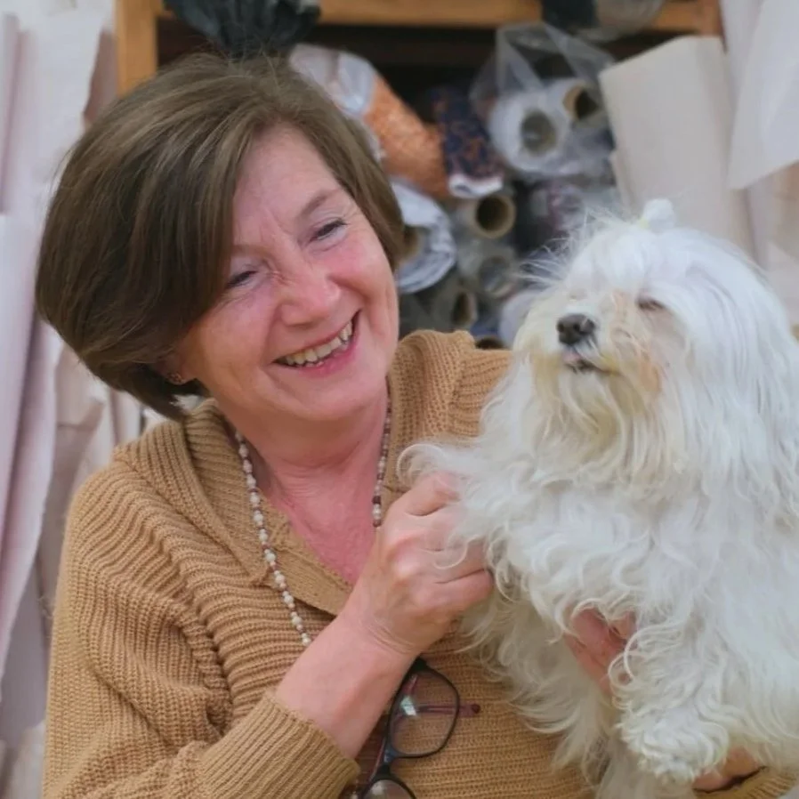A woman with short brown hair smiling and holding a small white fluffy dog, with a background of fabric rolls and sewing supplies.