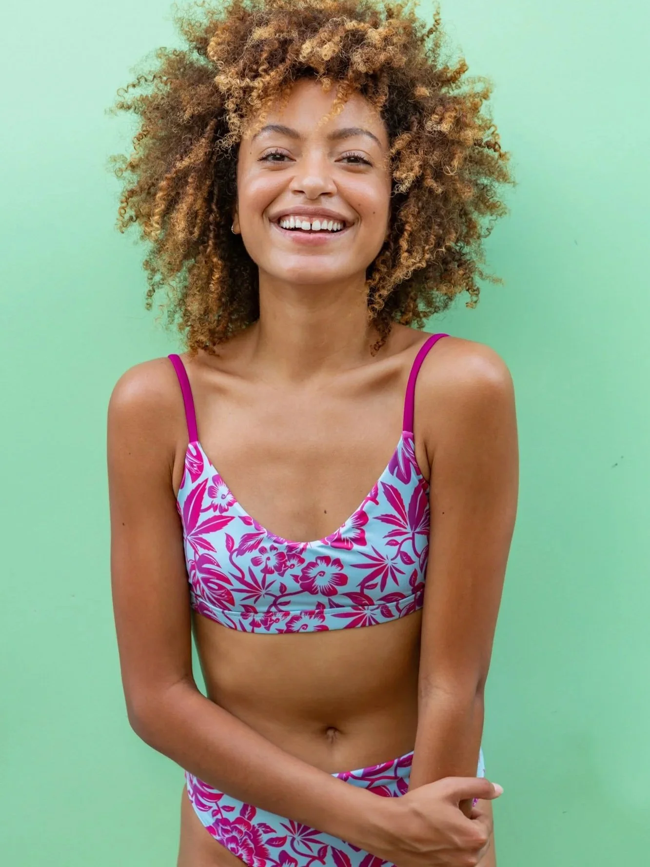 A young woman with curly hair wearing a floral bikini stands against a light green background, smiling at the camera.
