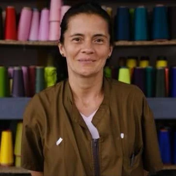 A woman standing in front of shelves filled with colorful spools of thread, wearing a brown jacket.