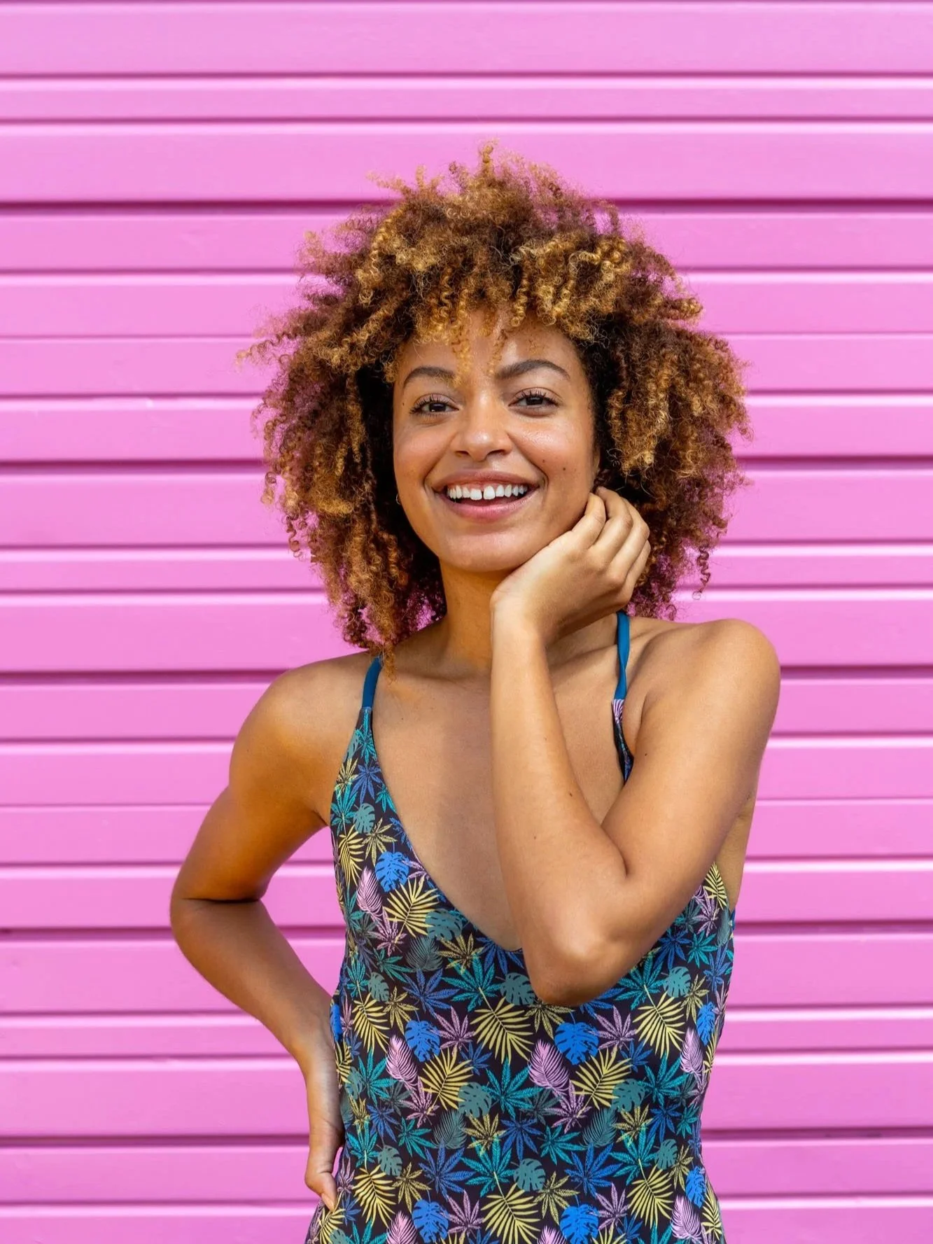 A young woman with curly, blonde hair standing in front of a pink wall, smiling, wearing a colorful sleeveless dress with a tropical leaf pattern.