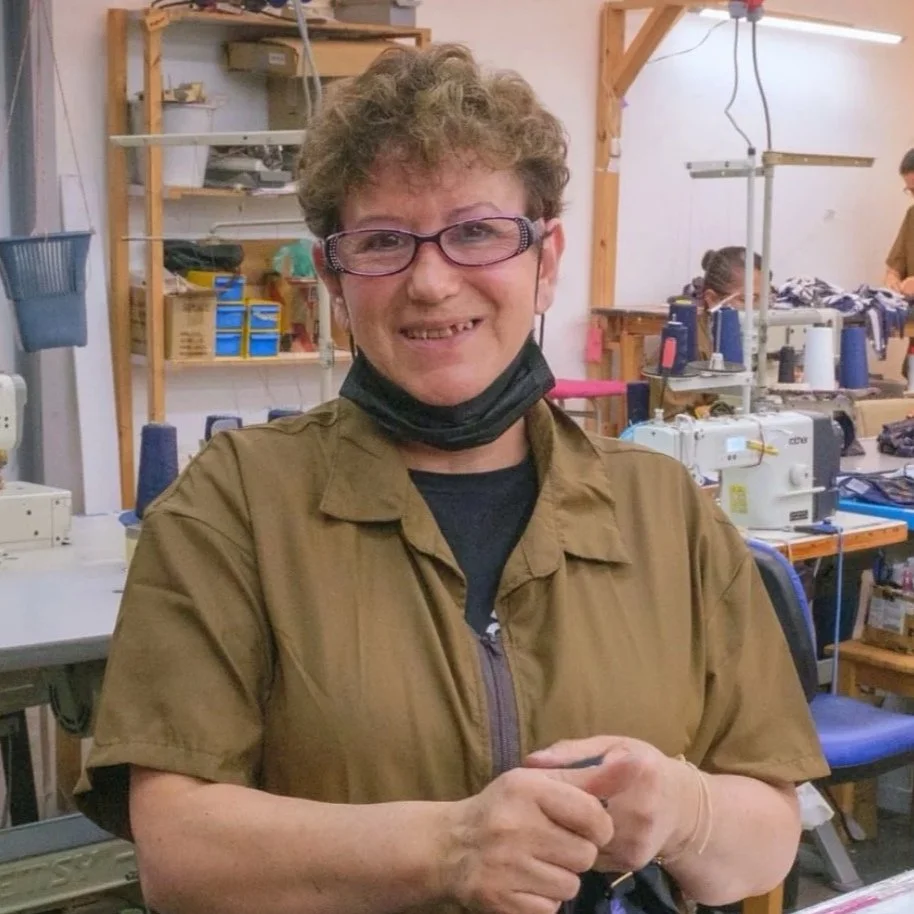 A woman with curly short hair, glasses, and a black face mask around her neck, smiling and holding her hands together, standing in a sewing or craft workshop.