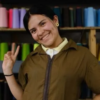 Young woman with short black hair smiling and making a peace sign with her right hand, standing in front of shelves filled with colorful fabric or paper rolls.