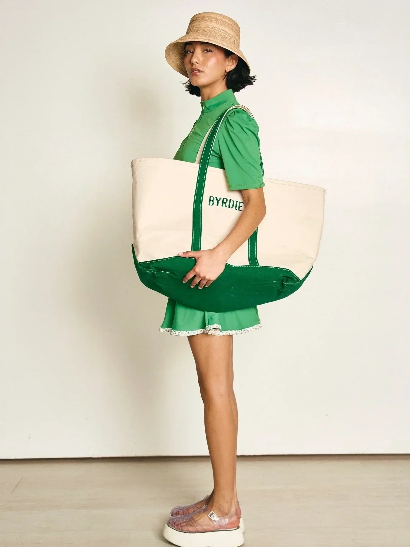 A woman wearing a tan wide-brim hat, green short-sleeved shirt, a green skirt with lace trim, light pink shoes, and holding a large beige and green tote bag with the word 'BYRDIE' on it, standing against a plain off-white background.
