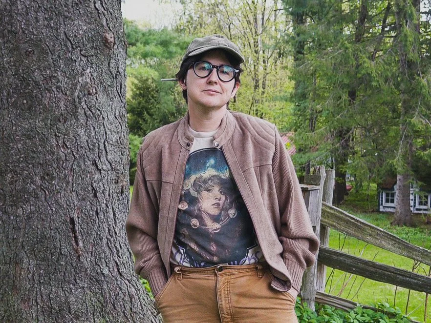 White guy in a brown suede jacket, baseball cap, and Stevie Nicks t-shirt. He's got glasses.