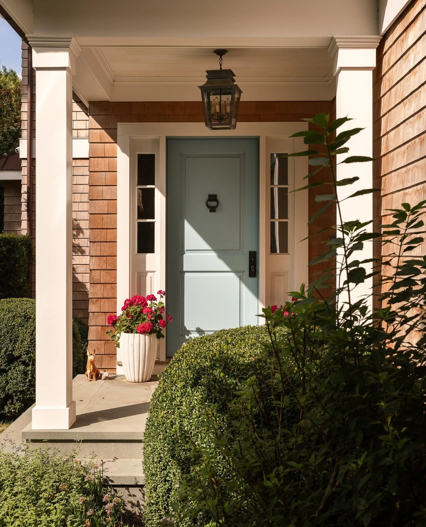 Afternoon light warms the cedar shingles and soft blue door of the front portico&mdash;a simple yet welcoming moment that captures the home&rsquo;s coastal spirit. The architecture balances traditional proportions with a clean, modern sensibility, se