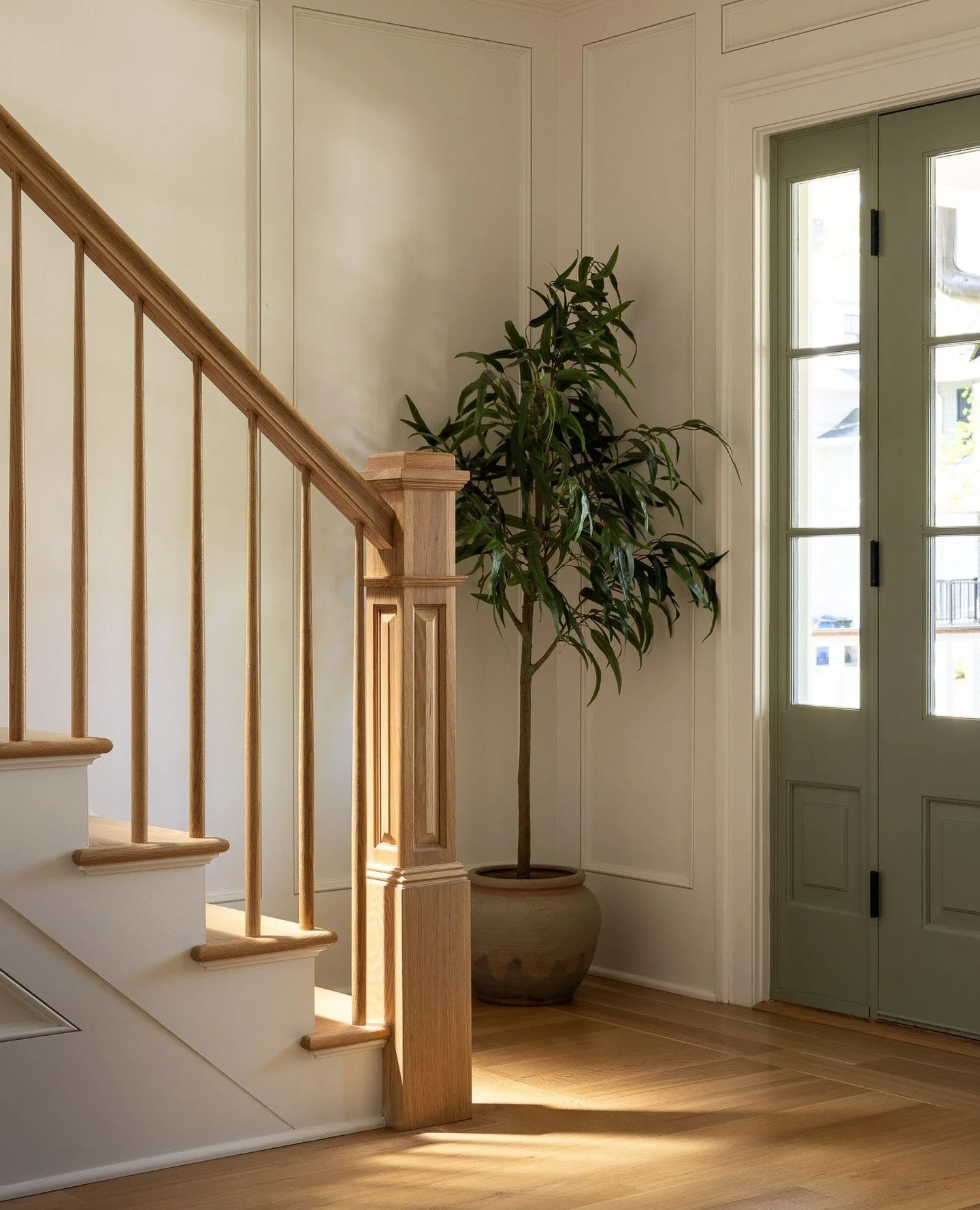 The new paneled foyer is washed in natural light, where a sage-green front door and oak stair rail create a welcoming entry. Though not visible here, a stained-glass window nearby casts dramatic light patterns into the space throughout the day, enric