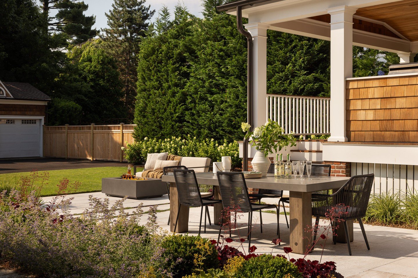 Just beyond the covered porch, a new open patio extends the rhythm of daily life outdoors. A dining terrace anchors the space, while a firepit lounge sits in the distance for evenings by the fire. A cedar fence continues the warm, natural material pa