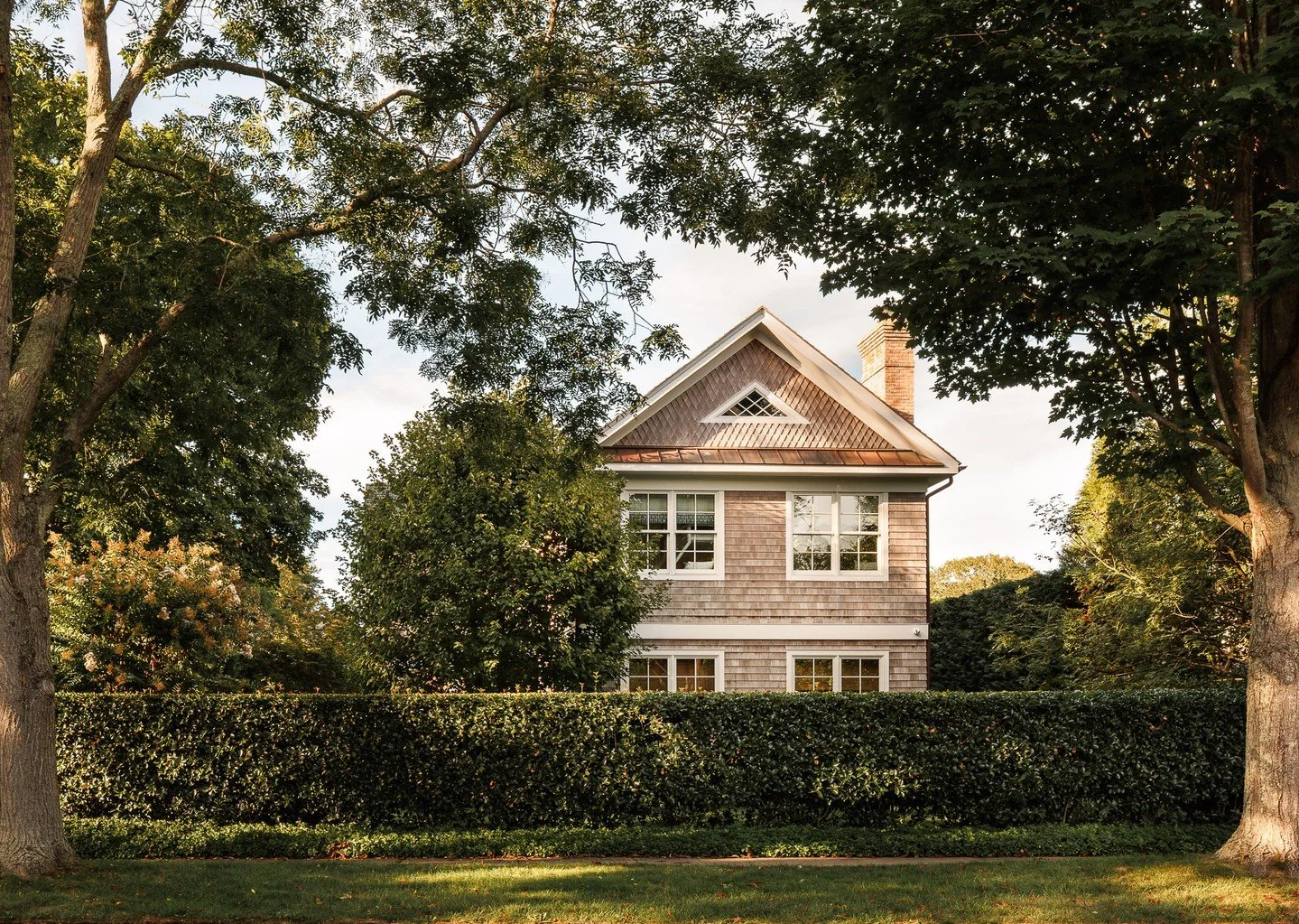 Set back behind a clipped hedge and framed by mature trees, the home presents a restrained cedar fa&ccedil;ade with a gabled roofline and standing seam accents. The architecture nods to Hamptons tradition while concealing a highly efficient, light-fi