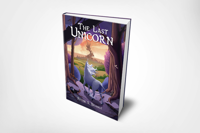  A book standing vertically on a white field. The book is “The Last Unicorn” by Peter S. Beagle. The cover shows a unicorn looking out of the lilac wood towards a crumbling castle on  the horizon.   