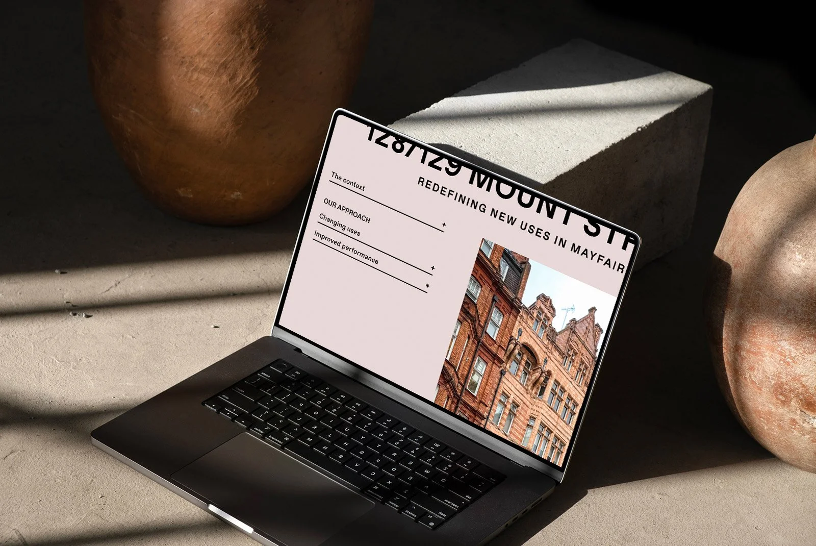 Laptop displaying a presentation slide titled 'Redefining New Uses in Mayfair' with sections on 'The context,' 'Our approach,' and 'Improved performance,' set on a concrete surface among large decorative pots and stones.