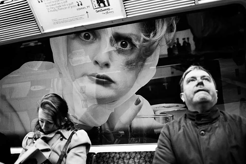 Black and white photograph showing a street scene with two people looking upward at an oversized close-up portrait of a face displayed on a billboard or storefront.