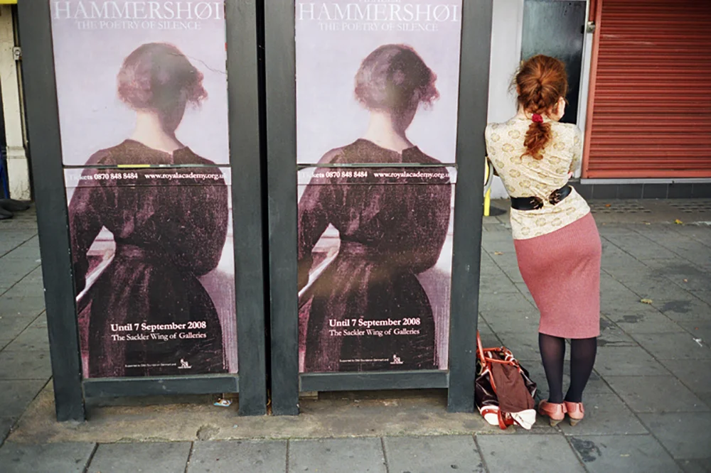 Street photography capturing a woman viewing two identical posters for a Hammershoi exhibition featuring a portrait of a figure from behind.
