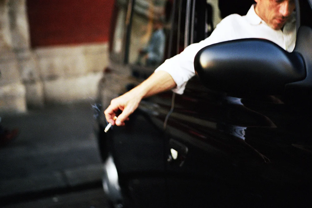 A colour film photograph of a person leaning out of a car window wearing a white shirt, holding a cigarette.