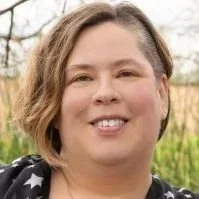 The face of Dr. Jo Eckler, licensed psychologist, a smiling nonbinary white person with short light brown hair cut asymmetrically standing in front of grass and trees.