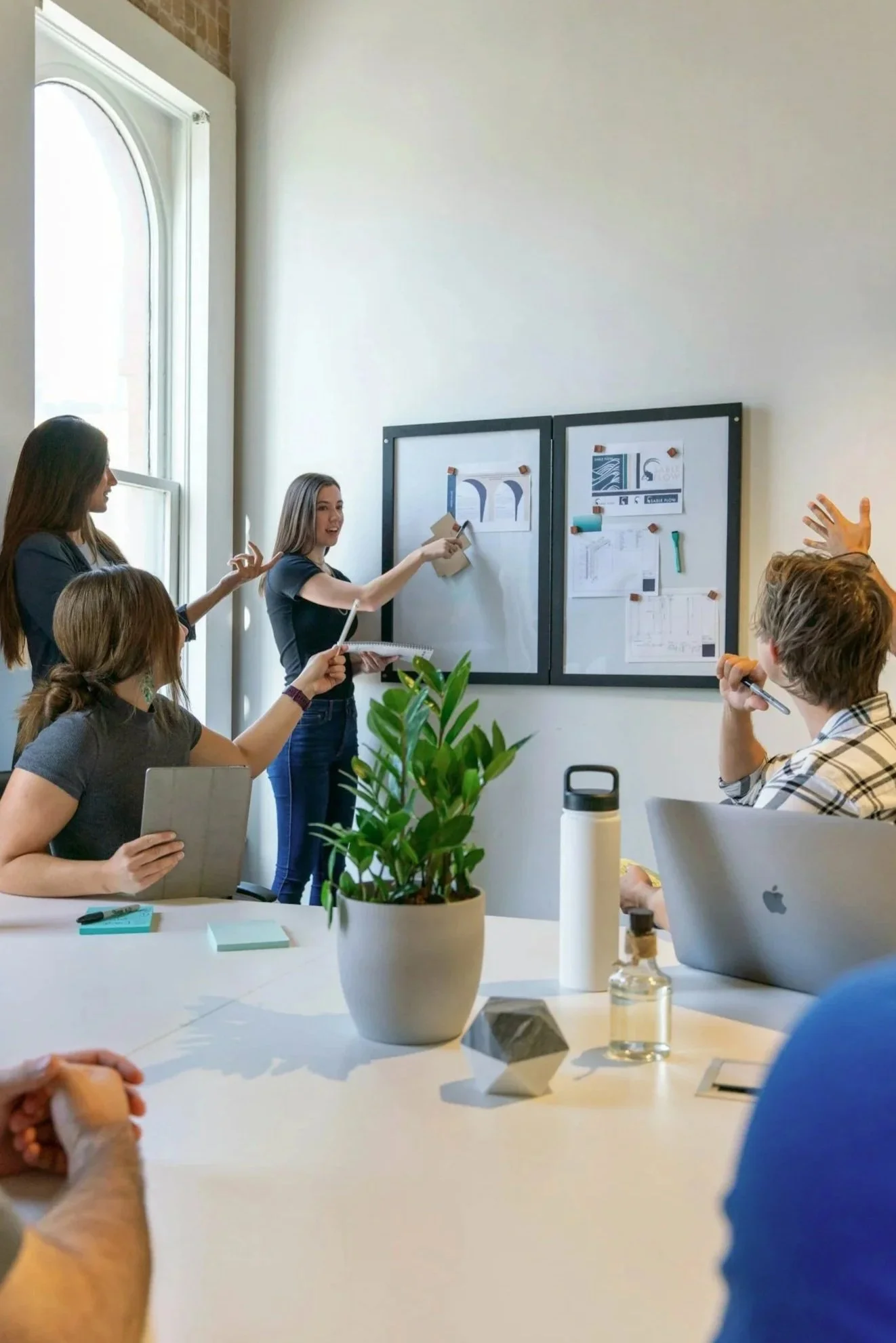 In a sunny meeting room, a woman stands at a board in the front of the room while people seated at the table make gestures as if they are sharing ideas.