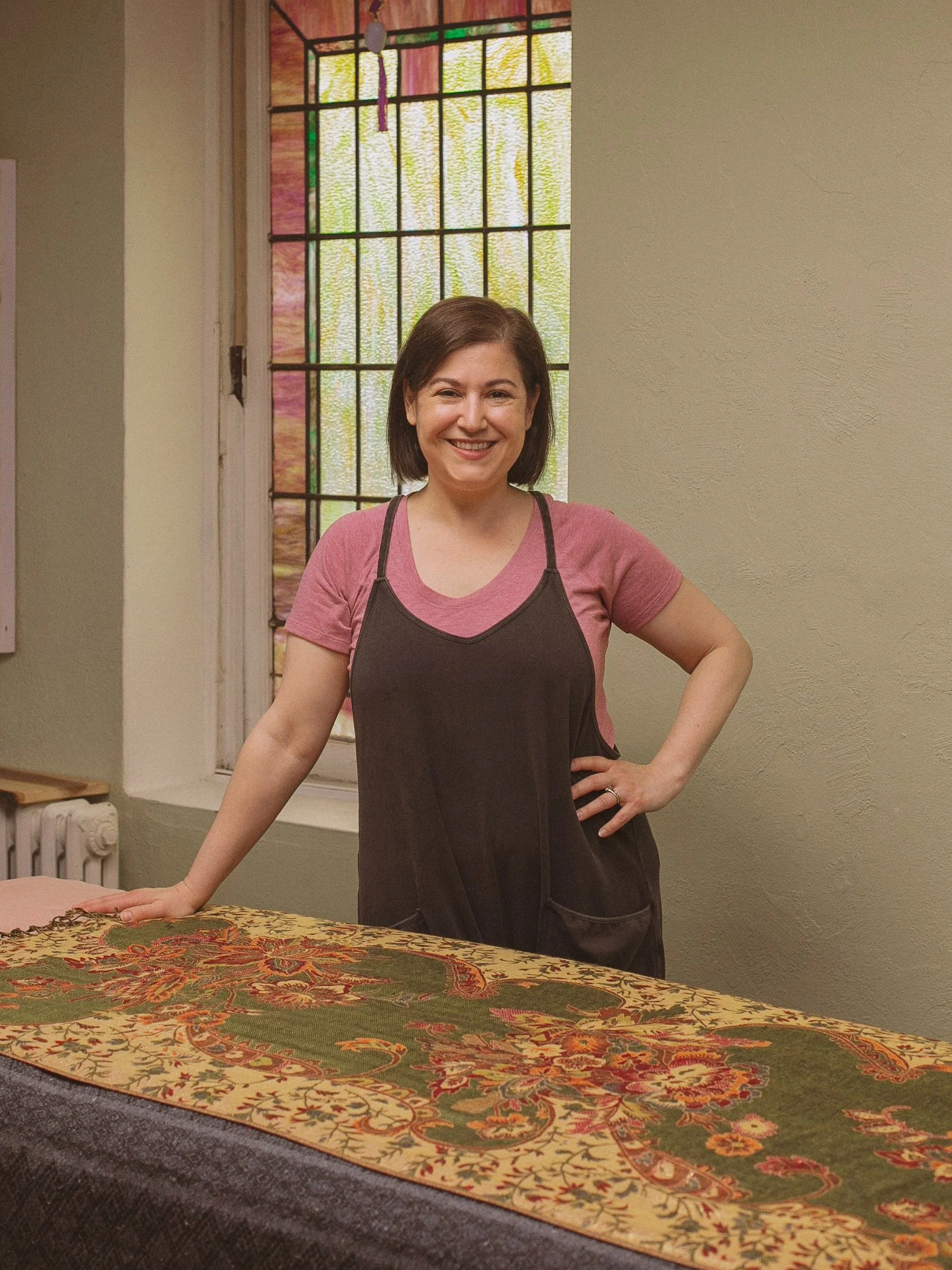 Kat Duvic smiling, standing in her studio with a large stained glass window, located inside of the Lake Harriet Spiritual Community.