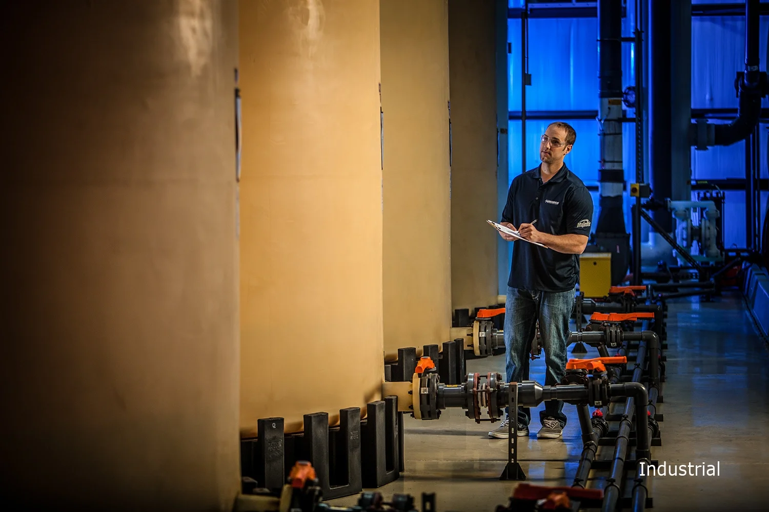 A man wearing glasses and a black polo shirt with logos, holding a clipboard and pen, inspecting industrial equipment with pipes and large tanks in the background, in an industrial setting with blue lighting.