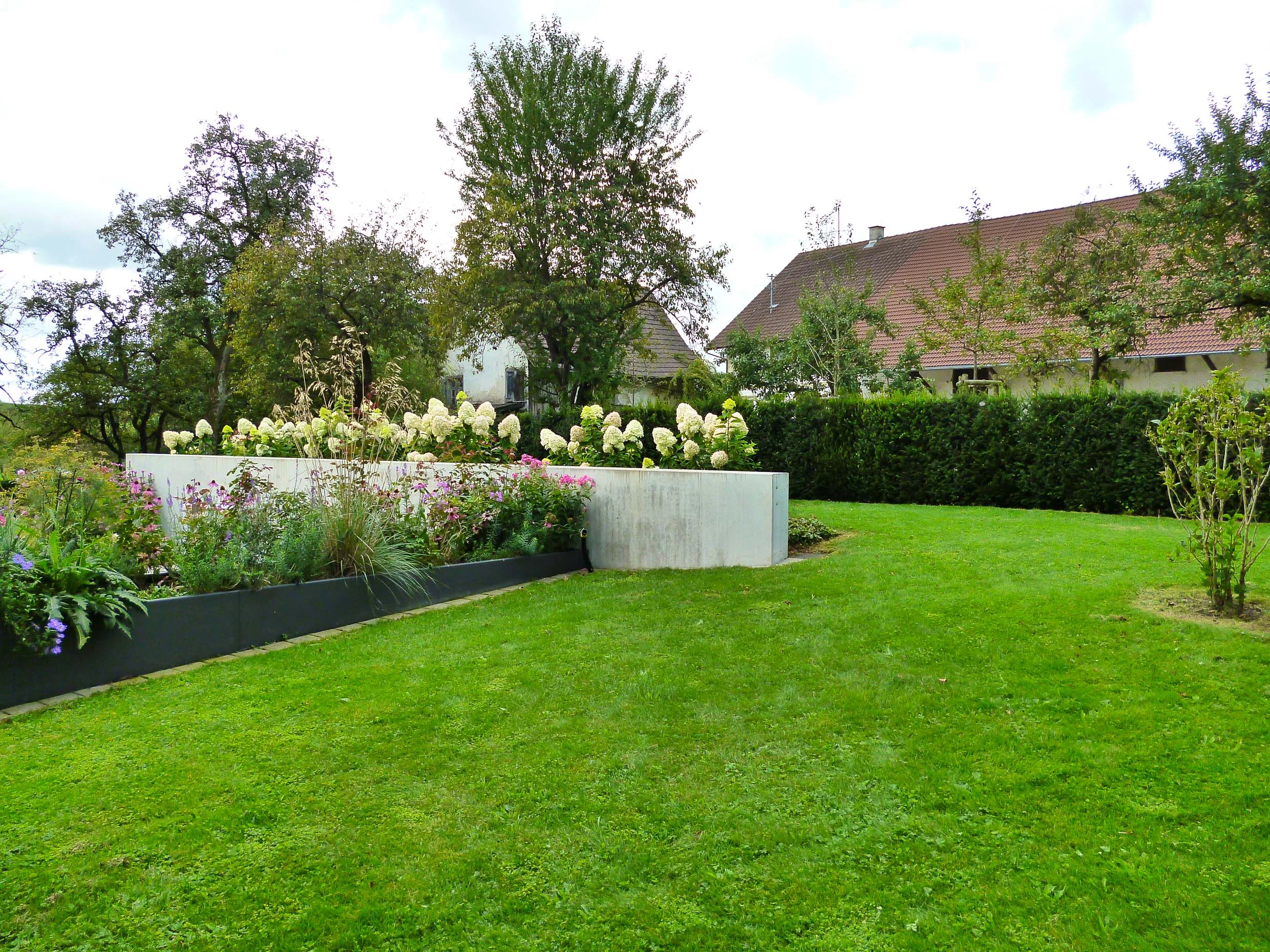 Die strenge Architektur setzt sich auch im Garten fort. Ein blühender Trog mit Rosen und Stauden umschließt einen großen Sitzplatz. Hinter der Stützmauer leuchtet eine Reihe von weißen Hortensien, die vor einer dunkelgrünen Eibenkulisse ins Szene gesetzt wird. Es handelt sich um ein großes Gelände in offener Lage. Um Effekte zu erzielen, braucht es größere Stückzahlen von einer Art. Weniger Vielfalt bei den Pflanzungen vereinfacht auch die Pflege sehr.