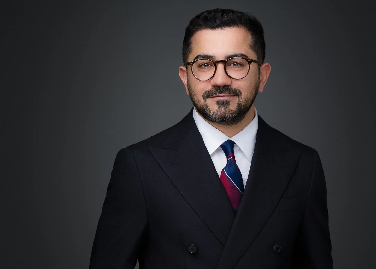 A man in a black suit, white shirt, and a striped tie, wearing glasses, posing against a dark background.