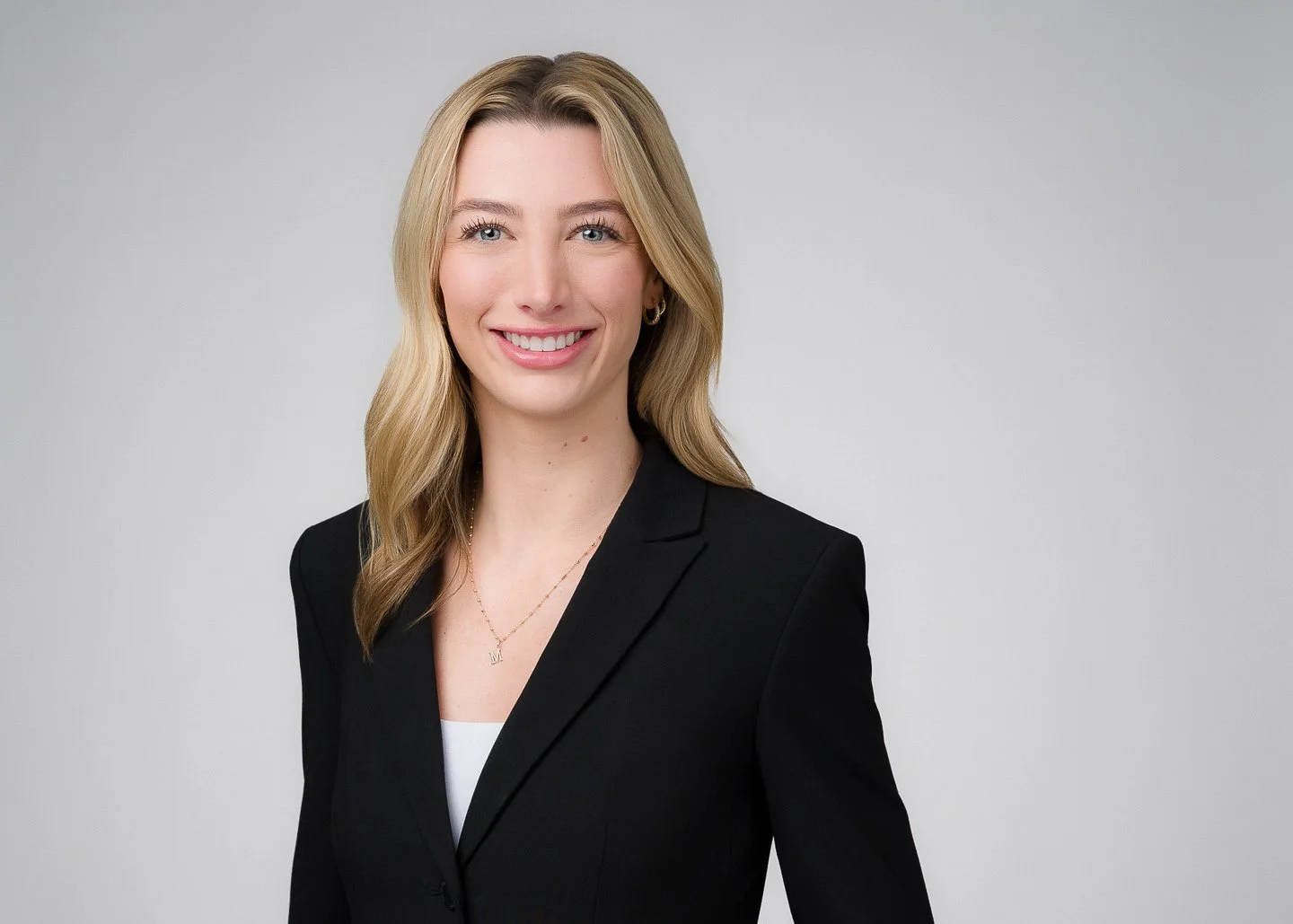 A professional woman with blonde hair, wearing a black blazer and a white top, smiling against a plain gray background.