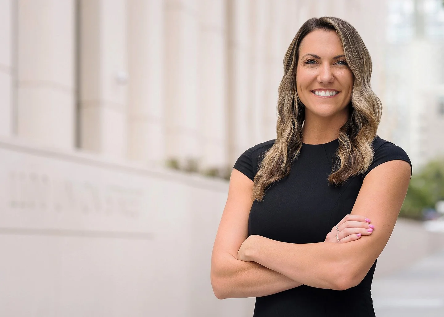 A smiling woman with wavy brown hair standing outdoors with arms crossed, wearing a black short-sleeved top.